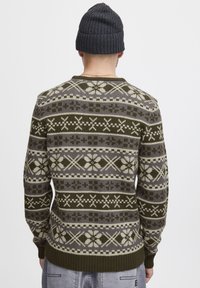 Strickpullover mit einem Muster aus Grün, Grau und Creme, das geometrische und schneeflockenartige Designs aufweist. Rippbündchen und Saum. Getragen mit einer dunklen Mütze.