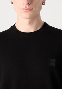 Zwarte gebreide trui met een ronde hals, voorzien van een klein vierkant stoffen label met de tekst "BOSS" op de linkerkant van de borst.
