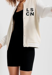 Jasno beżowa kurtka varsity z czarnymi literami "LSCN", długimi białymi rękawami i paskowanymi mankietami. Noszona na czarną sukienkę bodycon.