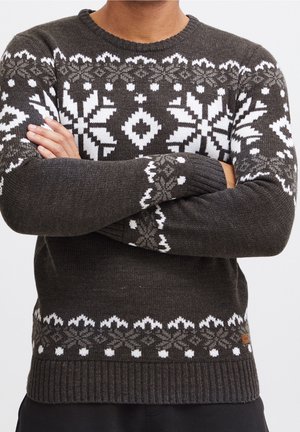 Homme portant un pull en maille gris foncé avec des motifs de flocons de neige et géométriques en blanc et gris clair, les bras croisés devant lui.