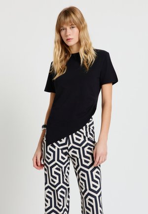 Donna con capelli biondi fino alle spalle indossa una maglia nera asimmetrica e pantaloni con motivo geometrico bianco e nero su uno sfondo semplice.