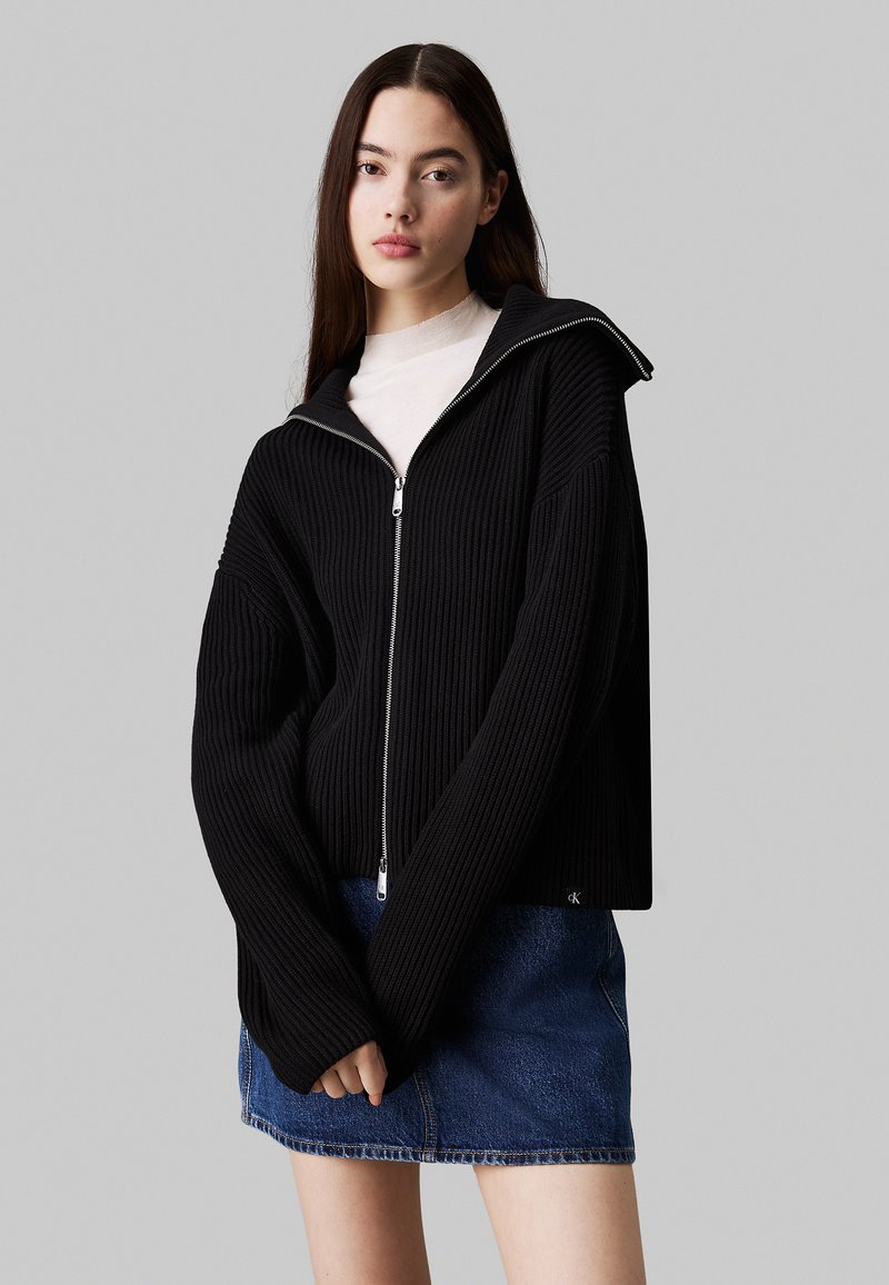Schwarze gerippte Zip-Hoodie mit hohem Kragen und übergroßen Ärmeln, getragen über einem hellen Oberteil und einem Jeansrock.
