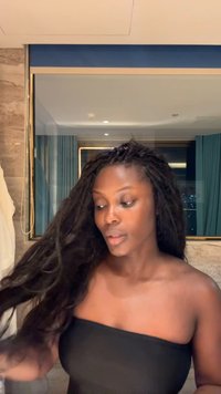 Kvinde med langt mørkt hår iført en sort top uden stropper, ser til siden i et badeværelse med et spejl og blå gardiner bag sig.