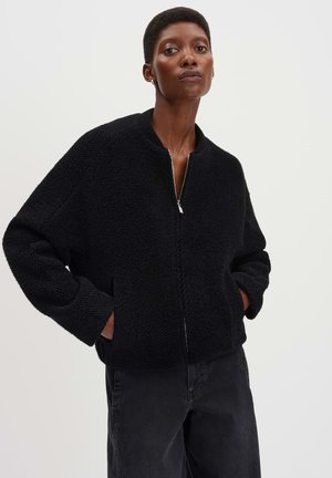Veste bomber noire texturée avec un col montant et une fermeture éclair intégrale. Présente des manches larges et des poches latérales. Portée sur un pantalon sombre.