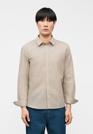 Młody mężczyzna w beżowej koszuli na guziki z delikatnymi paskami i niebieskich dżinsach, stojący na tle jednolitego białego tła.