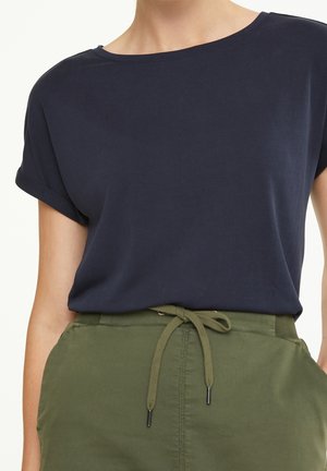 Vrouw draagt een navyblauw T-shirt met korte mouwen, ingestopt in olijfgroene high-waisted broek met taillekoord.