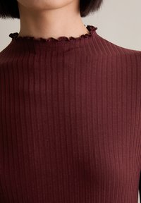 Burgunderfarbenes, geripptes Oberteil mit hohem Ausschnitt und gewelltem Saum, aus glattem, dehnbarem Stoff mit vertikalen Linien auf der Oberfläche.