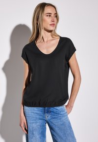 Svart blus med kort ärm och rundad halsringning, elastisk nederdel och slät tyg. Bärs med ljusblå denimjeans.