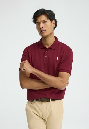 Hombre con los brazos cruzados, vestido con una camisa polo color burdeos y pantalones beige, de pie frente a un fondo claro y liso.