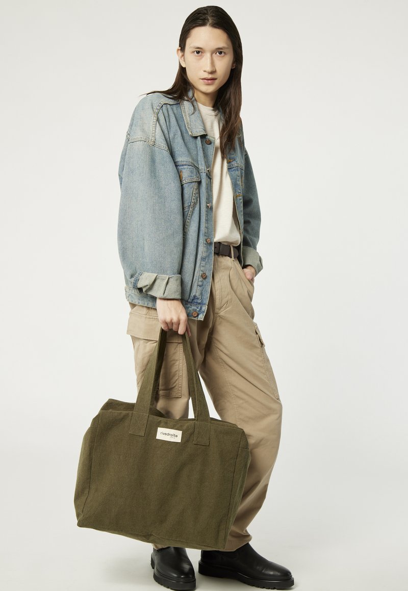 Sac fourre-tout en toile vert olive avec des poignées solides et une petite étiquette, associé à une veste en jean et un pantalon cargo beige.