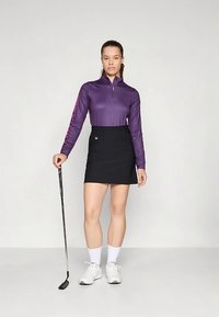 Γυναίκα που στέκεται με γκολφ κλαμπ, φορώντας μωβ μπλούζα με μακρύ μανίκι, μαύρη φούστα, λευκές κάλτσες και λευκά αθλητικά παπούτσια σε απλό φόντο.