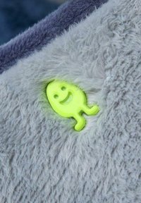 Tejido de felpa gris con una textura suave presenta un pequeño personaje sonriente en color verde lima adjunto, añadiendo un detalle juguetón.