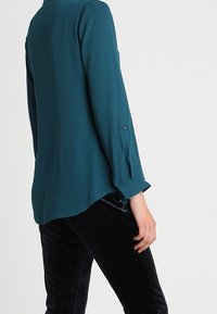 Teal, långärmad blus med knappade manschetter, lätt svängd nertill och mjukt tyg, parat med svarta plyschbyxor.