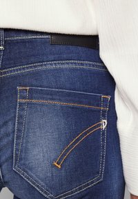 Jeans de mezclilla azul oscuro que muestran el bolsillo trasero con un detalle de costura naranja. Presenta una etiqueta en la cintura negra y una textura sutil.