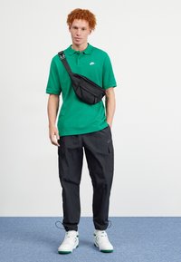 Vihreä polo-paita, mustat cargo-housut ja valkoiset tennarit vihreillä yksityiskohdilla. Musta crossbody-laukku, sileä kangas, rento istuvuus.