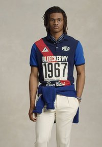 Sötétkék polosó piros és kék részletekkel, fehér "BLEECKER NY" és "1967" felirattal. Fehér nadrág és kék pulóver a derékon megkötve.