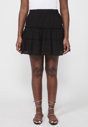 Jupe noire à volants avec motif ajouré, taille élastique, longueur courte et volants superposés. Associée à des sandales argentées à brides.