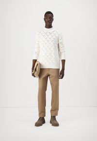 Maglione bianco lavorato a maglia con motivo a rombi, pantaloni beige e stivaletti marroni, tenuti con una borsa color nocciola. Design semplice su uno sfondo neutro.