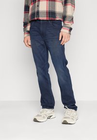 Jeans din denim albastru cu croială dreaptă, având un design clasic cu cinci buzunare. Purtați cu o cămașă în carouri și teniși de culoare deschisă.