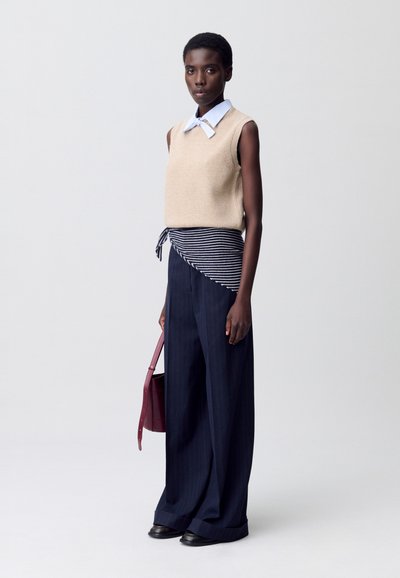 Maglione senza maniche beige sopra una camicia a righe blu e bianche, abbinato a pantaloni larghi blu navy. La modella tiene una borsa bordeaux.