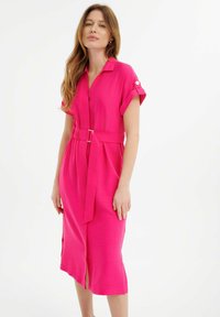 Abito rosa con cintura, colletto, maniche corte e spacchi laterali; il tessuto testurizzato aggiunge interesse visivo.