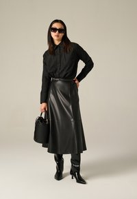 Chemise noire à boutons avec des détails perforés, jupe en cuir noir taille haute, bottes noires jusqu'aux genoux et sac seau noir texturé.