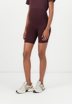 Person iført mørkelilla tætsiddende shorts og matchende top, stående med beige og hvide Nike-sneakers på en ensfarvet hvid baggrund.