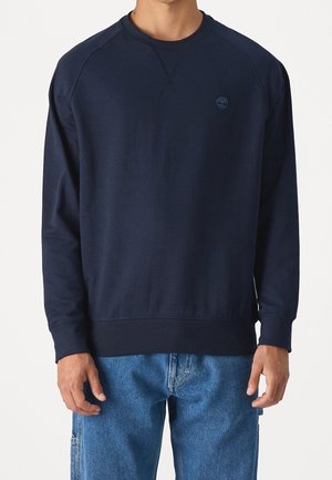 Mand iført en marineblå crewneck sweatshirt med et lille præget logo på venstre bryst og blå cowboybukser.
