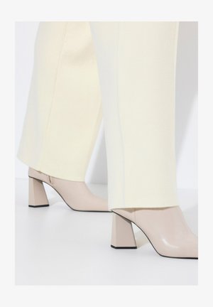 Beige korkokengät, joissa on tukeva korko, yhdistettynä kermaisiin, muotoiltuihin pantsuihin. Kengissä on kiiltävä pinta ja terävä varvasmalli.