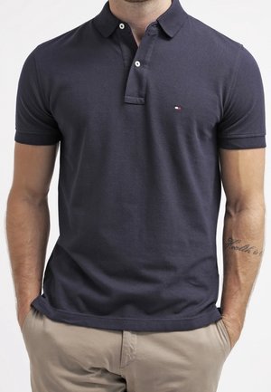 Homme portant un polo bleu marine foncé avec un petit logo sur la poitrine et un pantalon beige clair, les mains dans les poches.