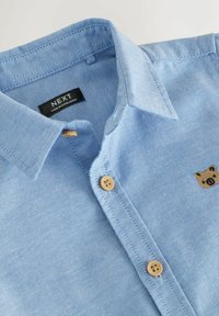 Cămașă bleu cu nasturi, confecționată dintr-un material texturat, având doi nasturi din lemn și un mic logo cu ursuleț pe piept.