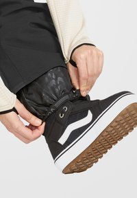 Schwarzer High-Top-Sneaker mit weißem Seitenstreifen, Obermaterial aus Stoff, Gummisohle und strukturiertem Grip. Fuß sichert die Hosenkante an dem Stiefel.