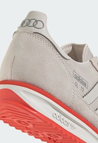 Baskets Adidas SL '72 avec une tige en suède gris clair, une semelle blanche et rouge, des anneaux argentés sur le talon et des accents à rayures.
