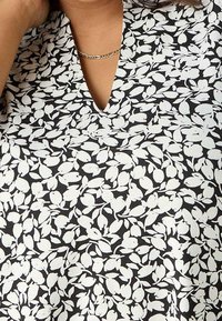 Primer plano de una persona que lleva una blusa negra con un patrón de hojas blancas y un collar de cadena dorada, mostrando el escote y parte del brazo.