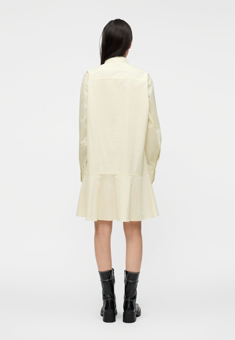 Robe chemise jaune clair avec un ourlet évasé, des manches longues et un col. Portée avec des bottines noires, présentant une texture lisse et une coupe ajustée.