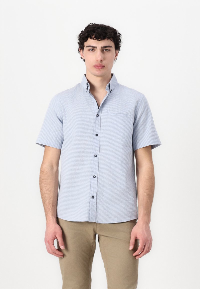 Camicia a maniche corte blu chiaro con un motivo testurizzato, chiusura con bottoni e taschino sul petto. Abbinata a pantaloni beige.