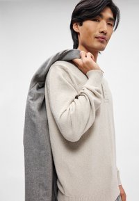 Pulover bej din tricot cu țesătură texturată și manșete cu nervuri, asortat cu o jachetă gri ușoară purtată pe umăr.