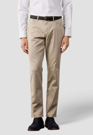 Hombre con pantalones beige, camisa blanca con botones, cinturón negro y zapatos de vestir negros, de pie frente a un fondo claro y liso.