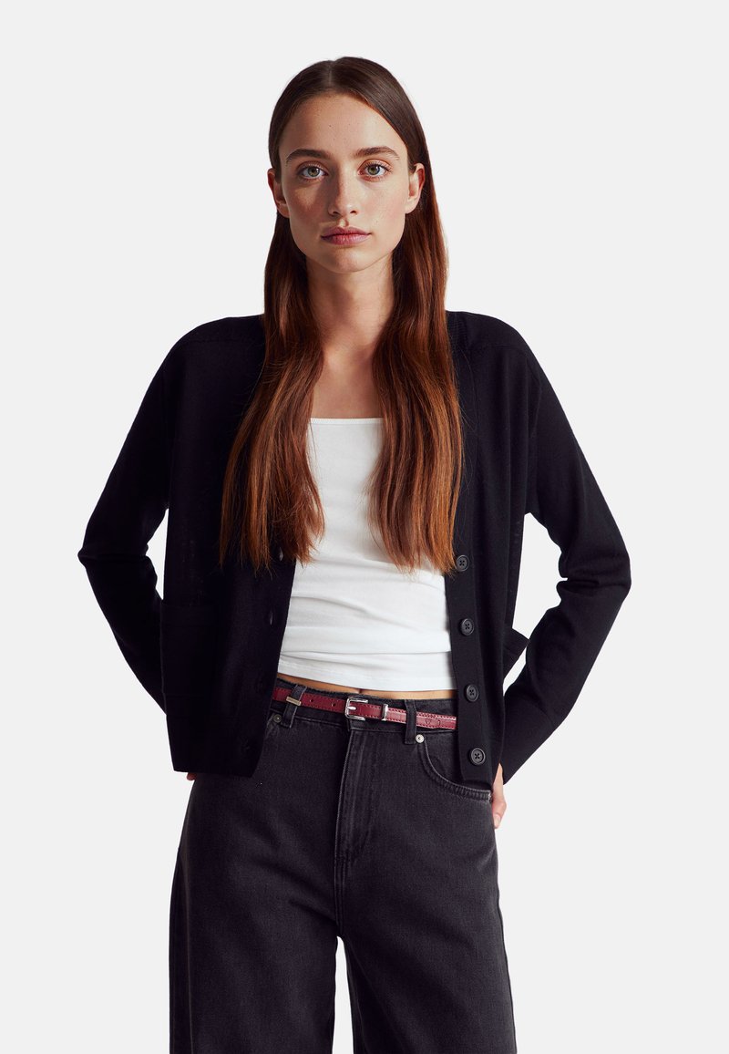 Cardigan nero con maniche lunghe, caratterizzato da una chiusura con bottoni e due tasche frontali, indossato sopra una maglietta bianca e jeans scuri.