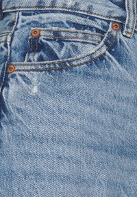 Ljusblå denimjeans har en sliten textur med fransiga områden, kopparnitar på fickor och kontrasterande gyllene sömmar.