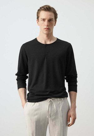 Jonge man in zwart henley shirt met lange mouwen en lichtbeige broek met trekkoord, staand met een neutrale uitdrukking tegen een effen achtergrond.