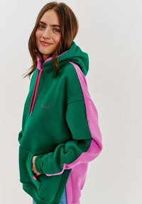 Veľká mikina s kapucňou so zeleným telom a ružovými rukávmi. Obsahuje šnúrky, prednú kapsu a malý vyšívaný detail. Vyrobená z mäkkej tkaniny.