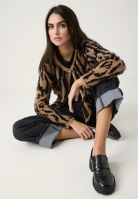 Flauschige braune und schwarze gemusterte Strickjacke mit Knopfverschluss, kombiniert mit schwarzen weit geschnittenen Hosen mit umgeschlagenen Säumen und glänzenden schwarzen Slippern.