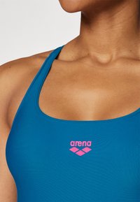 Plava kupaćica s izrezom i tankim trakama. Ime branda "arena" ispisano je u ružičastoj boji na području grudi. Glatka tekstura.