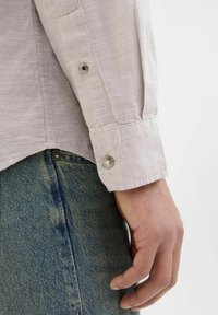 Camisa de manga larga color beige claro con un tejido texturizado, con puños abotonados y costuras visibles. Combinada con unos jeans de denim azul.