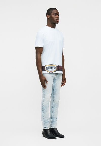 Άνδρας που φοράει ένα απλό λευκό μπλουζάκι με μεγάλο τύπωμα ζώνης, ανοιχτόχρωμα τζιν και μαύρες δερμάτινες μπότες, στέκεται μπροστά σε απλό φόντο.