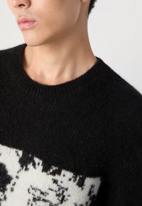 Black knit sweater with a round neckline, featuring a white abstract pattern at the hem; soft texture and thick knit fabric.