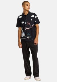 Schwarzes Shirt mit kurzen Ärmeln und einer Grafikdruck, der eine Figur und große weiße Flecken zeigt; kombiniert mit dunklen Jeans und weißen Sneakers.