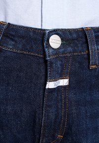 Vaqueros de mezclilla azul oscuro con un botón metálico visible etiquetado como "CLOSED", costuras naranjas y una etiqueta blanca en la cintura.