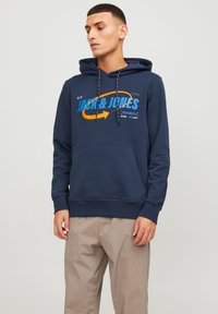 Sudadera azul marino con bolsillo frontal tipo canguro, cordón y texto gráfico. Fabricada en material suave, con acentos en azul y naranja contrastantes.